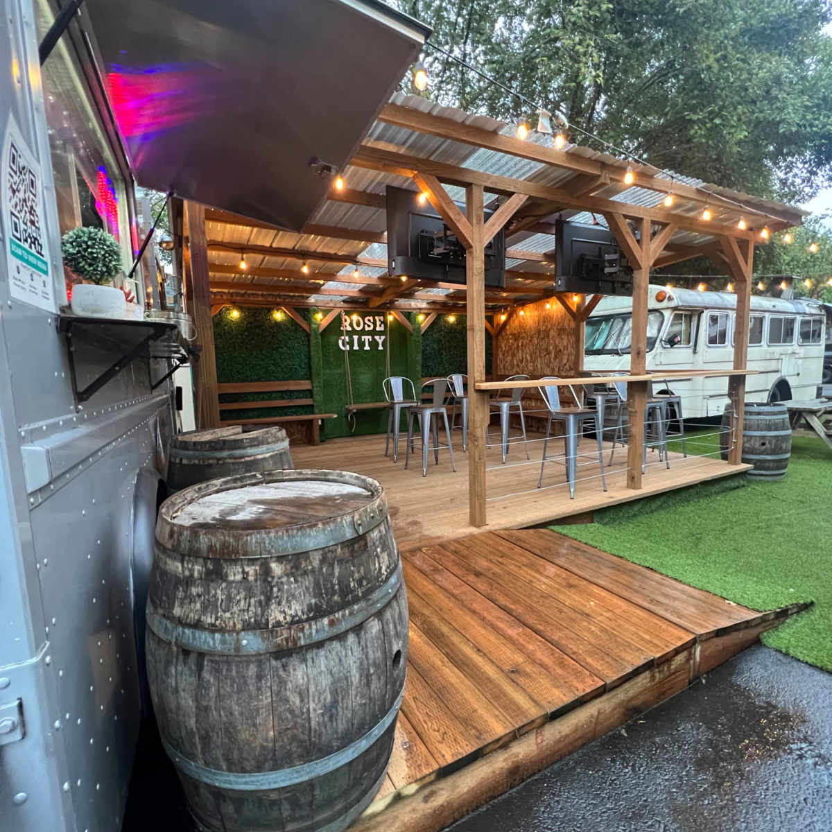 A food truck is positioned next to a wooden deck with floating lights and seating under a covered structure, surrounded by grass and other food trucks.