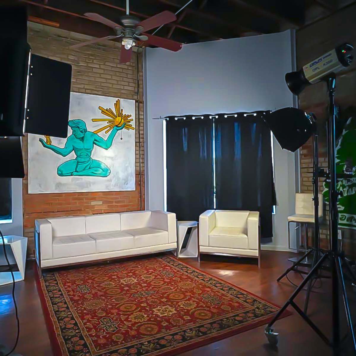 A well-lit studio space featuring a white sofa, a white armchair, a colorful wall painting, and photography lighting equipment.
