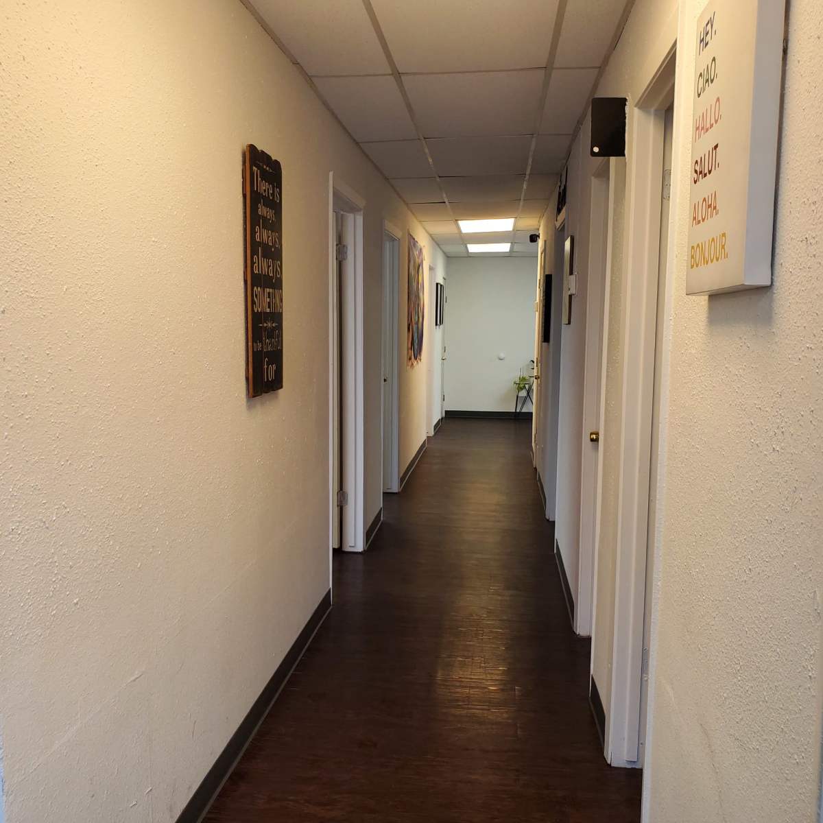 A long hallway with white walls, wooden flooring, and doors lining both sides, along with framed signs on the walls.