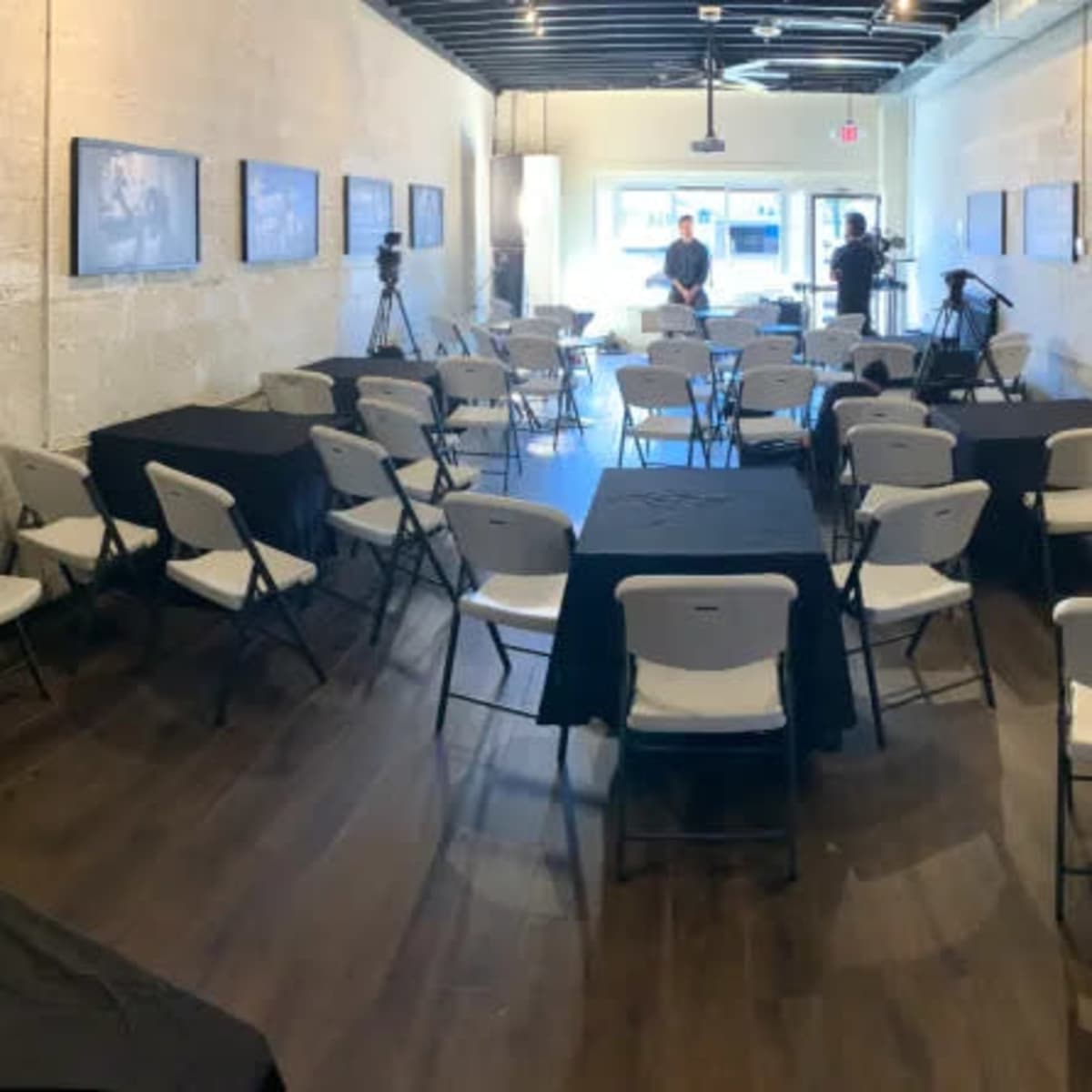 A spacious room with rows of white chairs and black tables arranged for an event, with photographs displayed on the walls.