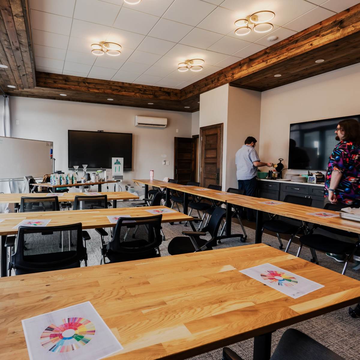 A modern meeting room features wooden tables arranged for a workshop, with colorful printed materials on each table and video equipment set up for presentations.