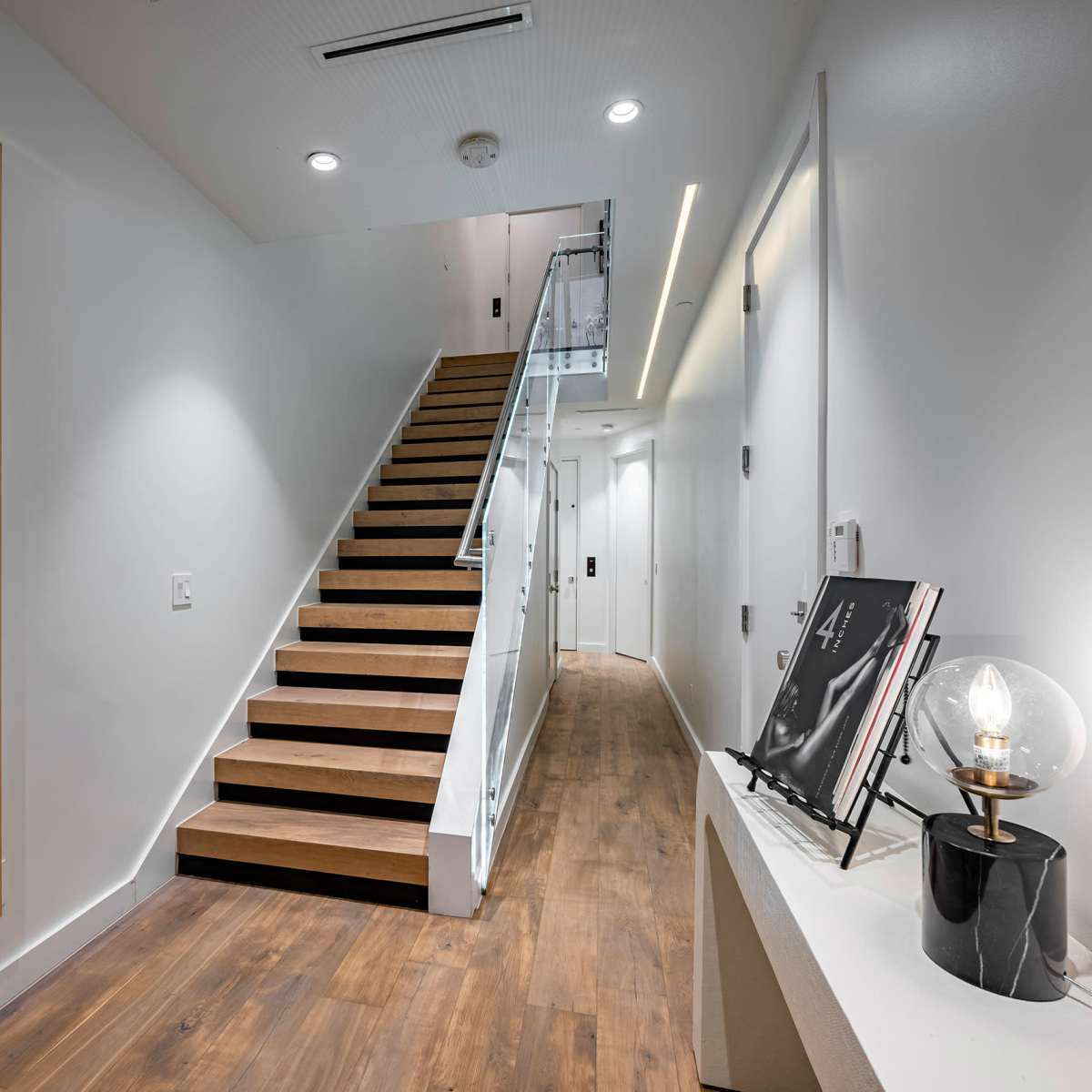 A modern hallway featuring a staircase with wooden steps, a large black and white artwork, and a small table with a lamp and a picture frame.