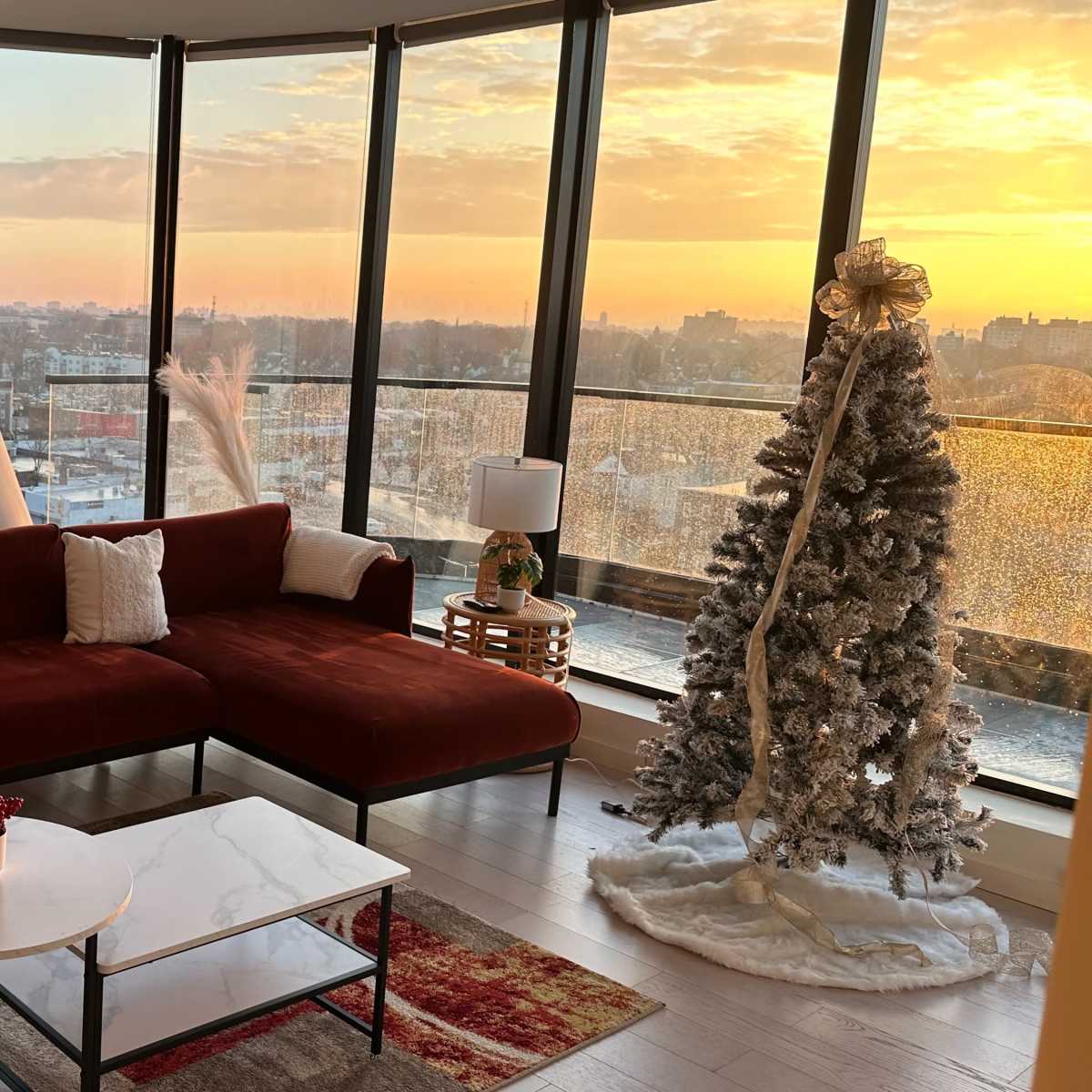 A modern living room features a large red sofa, a marble coffee table, and a decorated Christmas tree against a backdrop of floor-to-ceiling windows showcasing a sunset view.