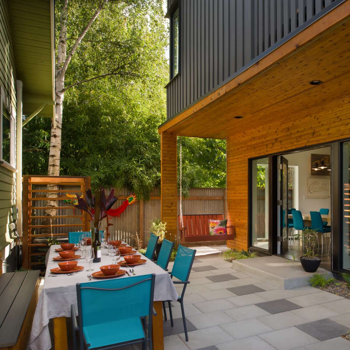 An outdoor dining area with a long table set for a meal is adjacent to a modern house featuring large glass doors and wooden elements.