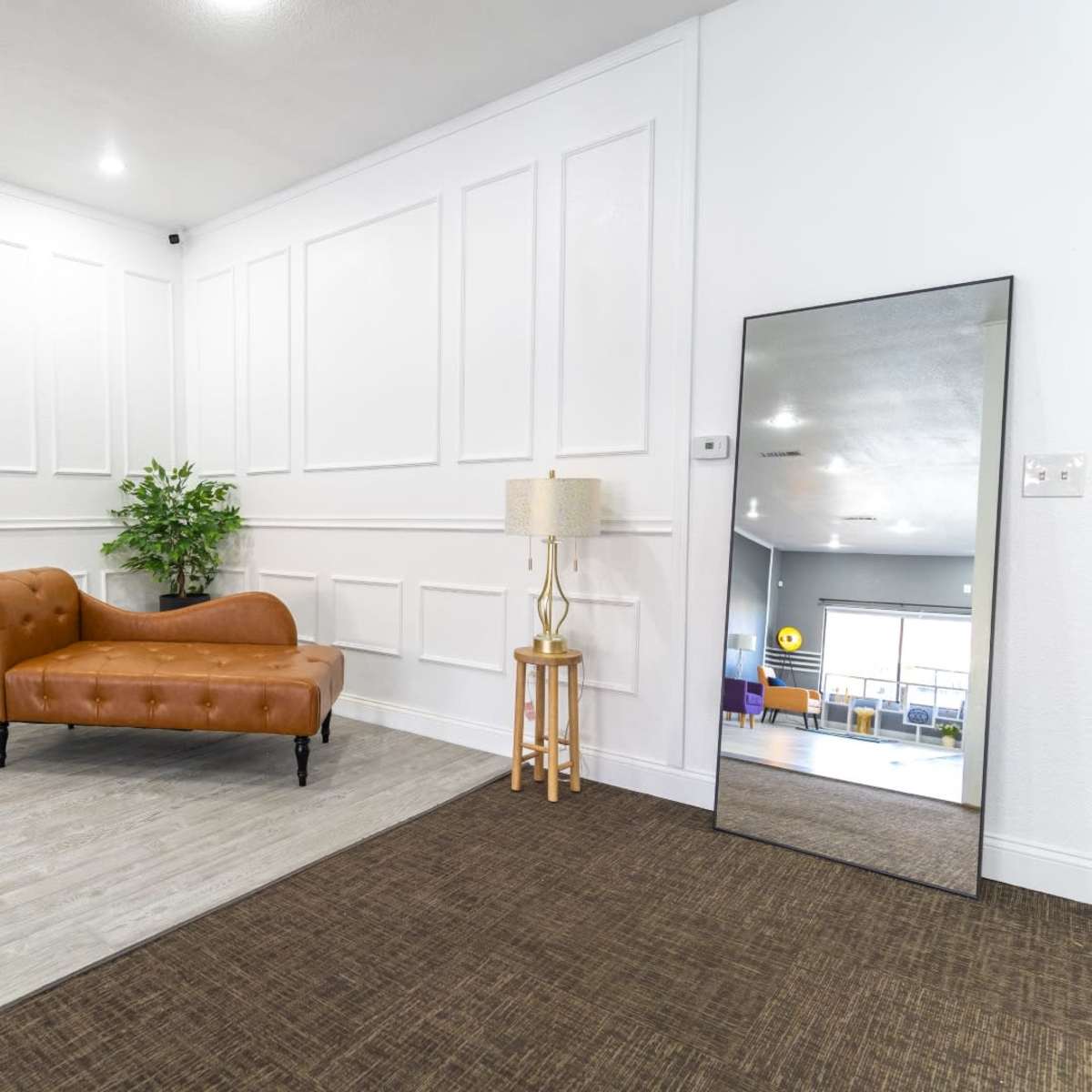 A room with a brown leather sofa, a decorative plant, a standing lamp, and a large mirror against a wall with paneling, along with a textured carpet floor.
