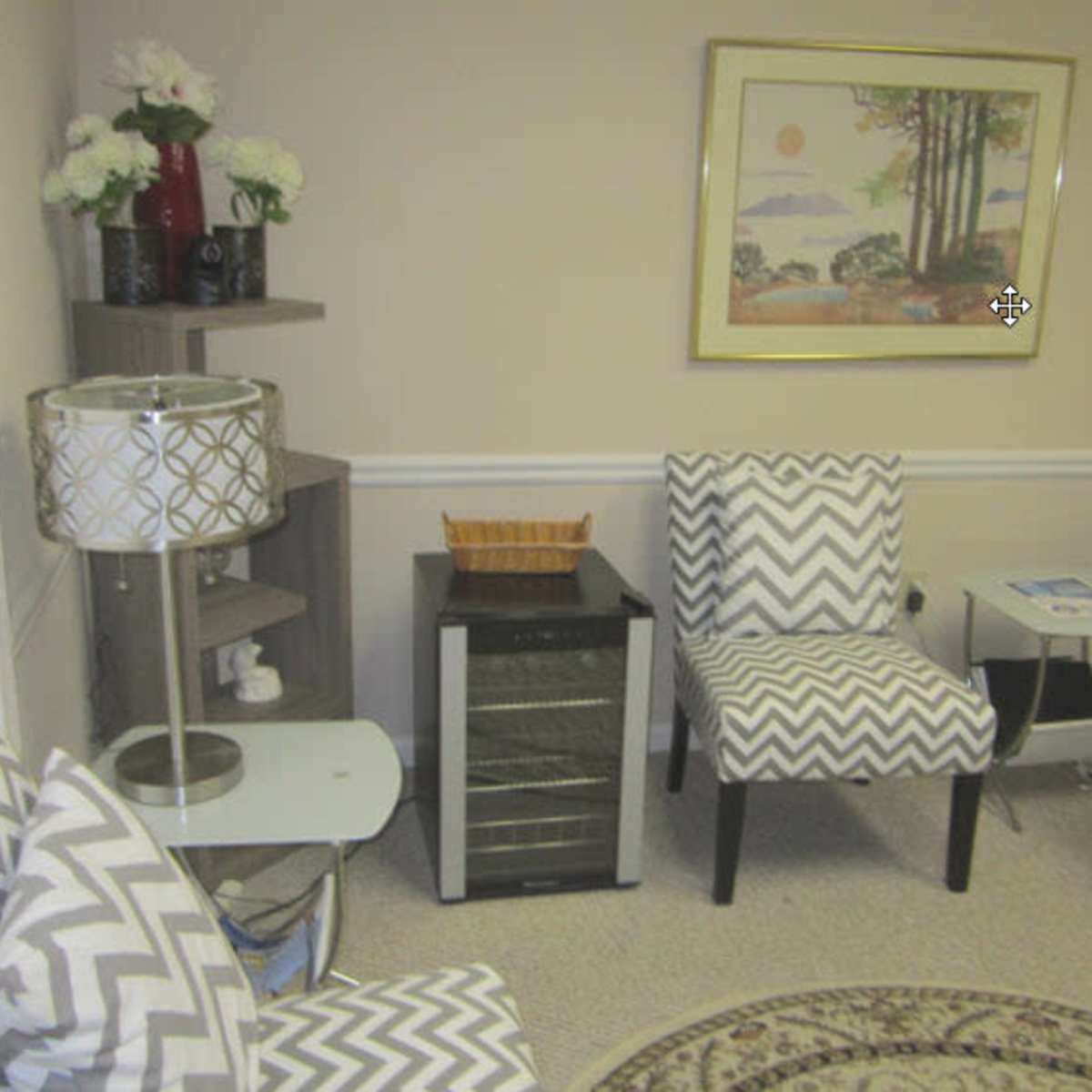 A small living area featuring two patterned chairs, a side table with a lamp, a wine cooler, and a decorative wall art piece.
