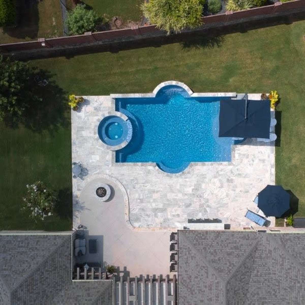 A backyard with a swimming pool featuring a hot tub, surrounded by a stone patio and lawn, with a playset in the corner.