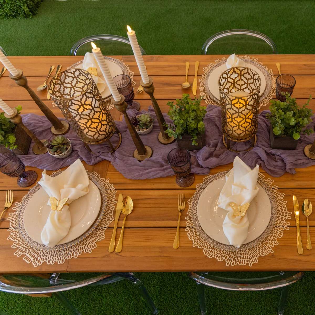 A long wooden table is set with ornate plates, gold cutlery, decorative napkins, and centerpieces featuring lanterns and greenery.
