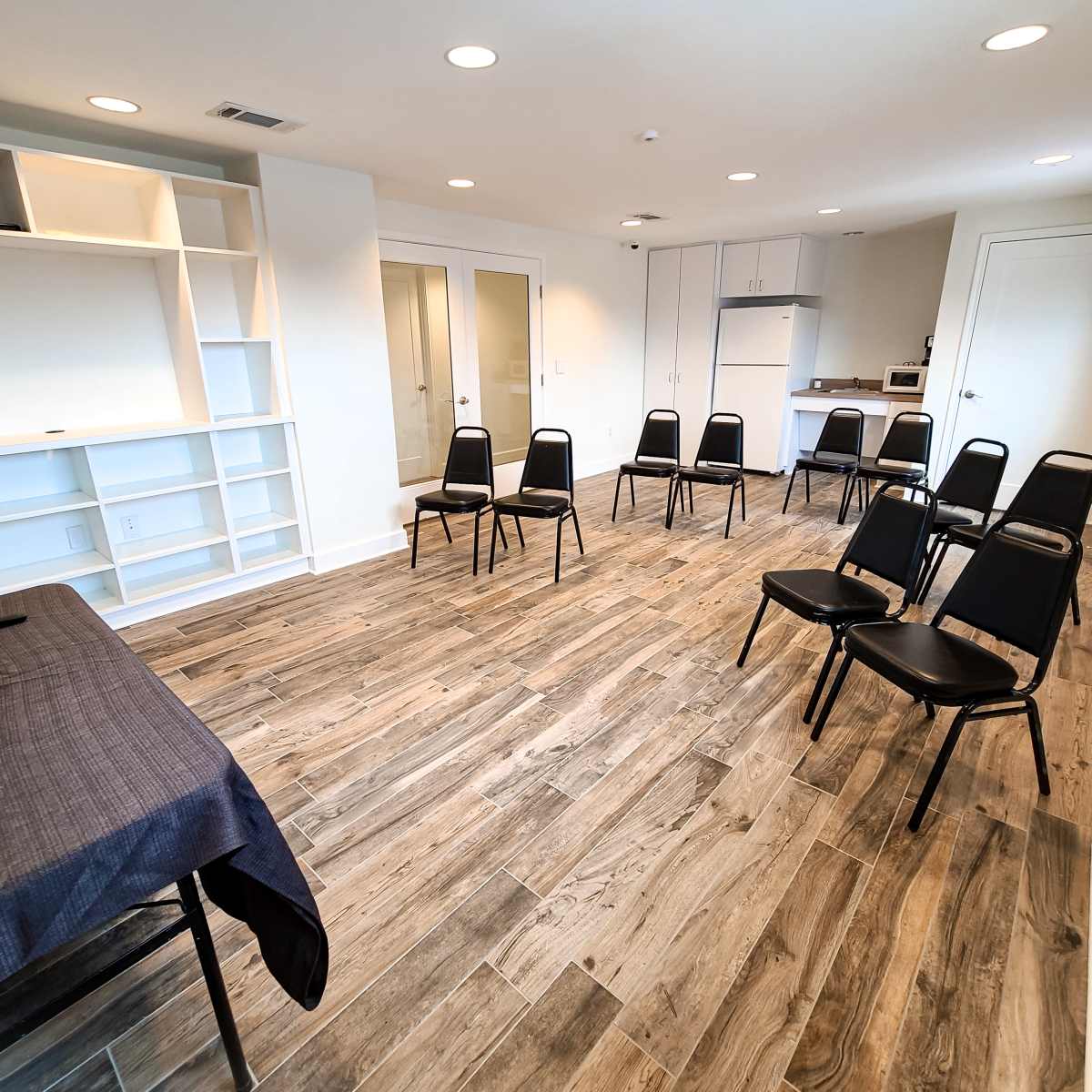 A modern meeting room with a table in front and several black chairs arranged in rows on a wooden floor.