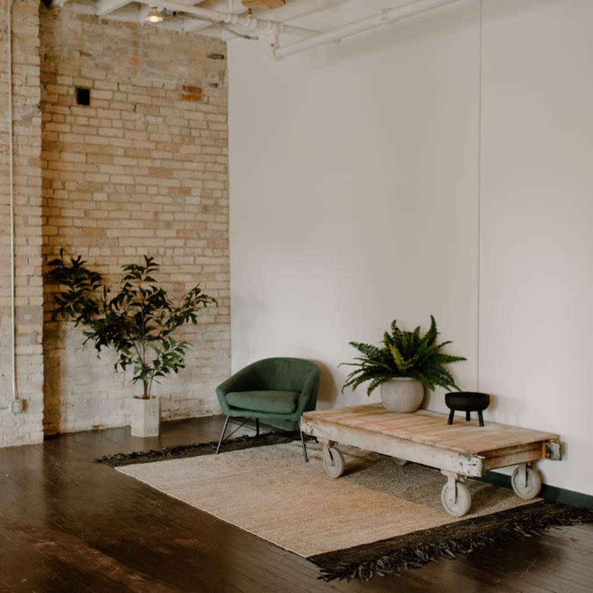 A minimalistic room with exposed brick walls, a green chair, a wooden table on wheels, and decorative plants on the floor.