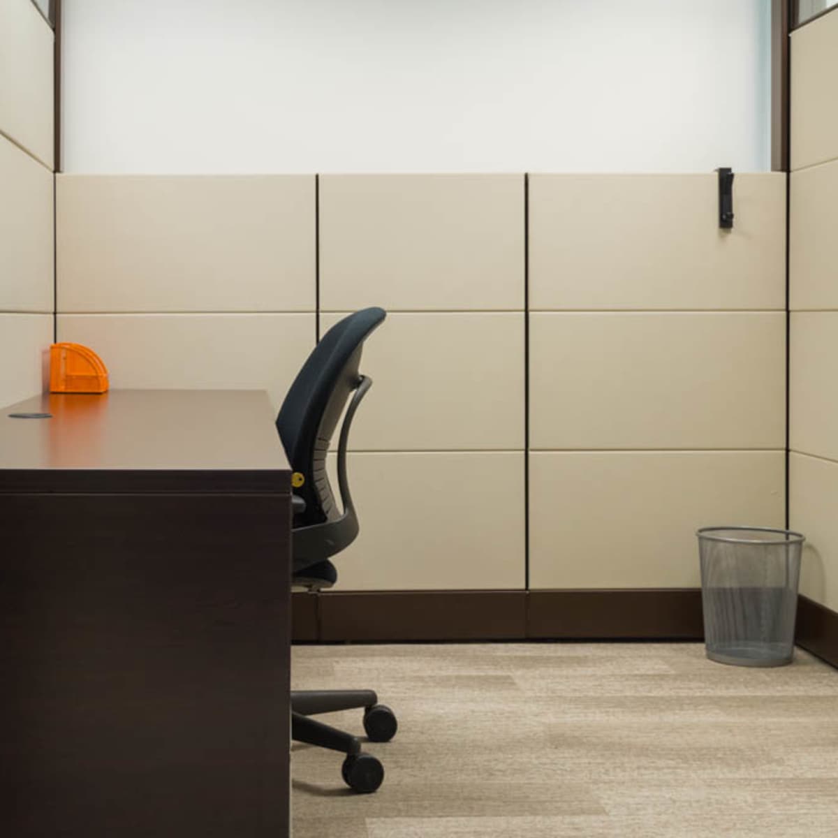 An empty office cubicle with a desk, an ergonomic chair, and a wastebasket in the corner.