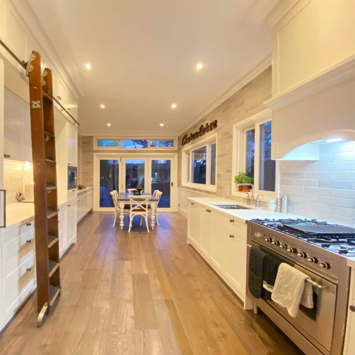 A modern kitchen features white cabinetry, wooden flooring, and a dining area with a table and chairs visible through large windows.