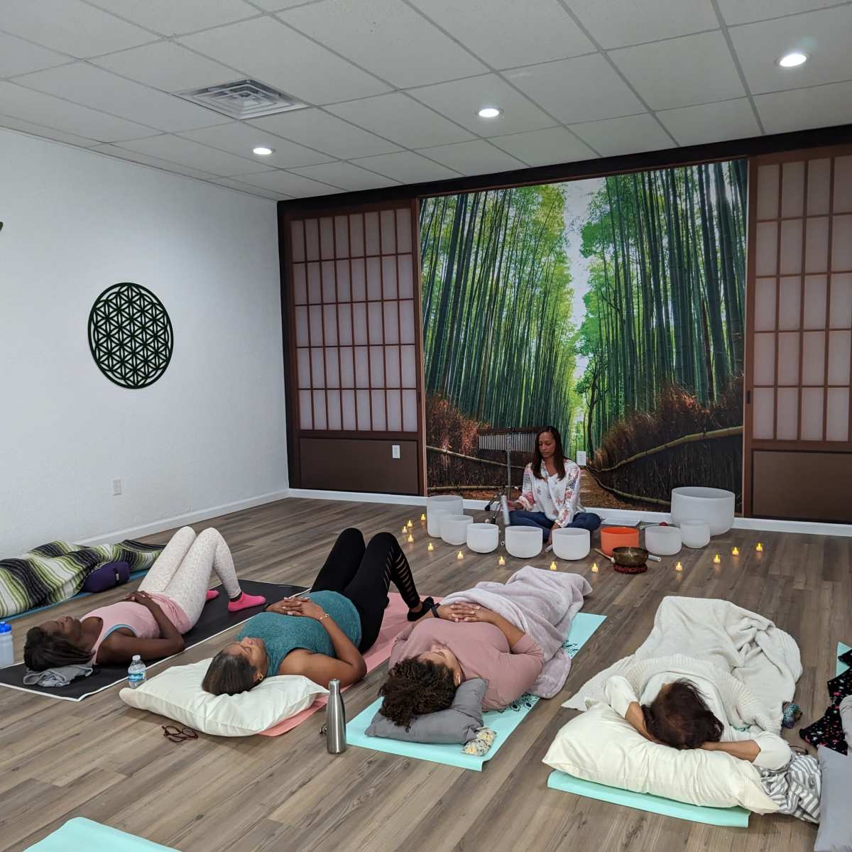 A group of individuals lies on mats in a yoga studio, while an instructor sits at the front with singing bowls in a serene setting adorned with a bamboo forest mural.