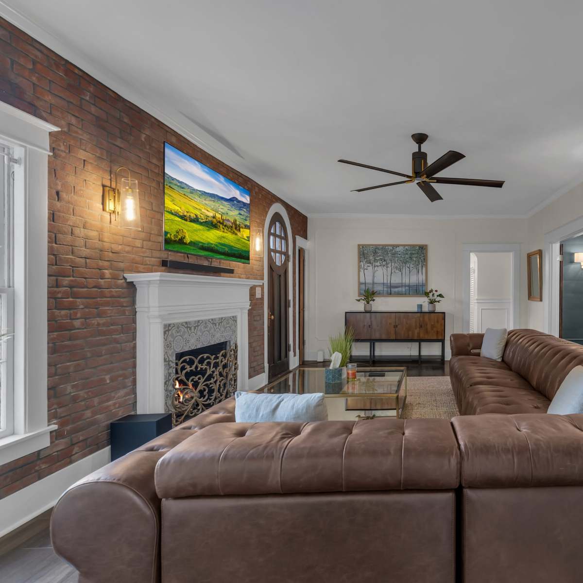 A spacious living room features a large brown leather sectional sofa, a brick accent wall, a mounted television, and artwork above a decorative fireplace.