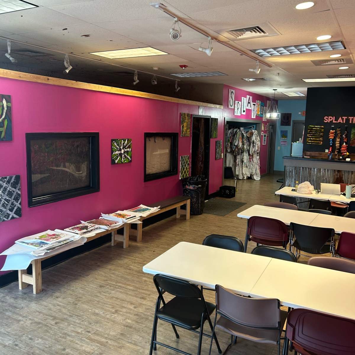 An art studio with bright pink walls, displaying artwork on the walls, tables set up for patrons, and separate areas for creative activities.