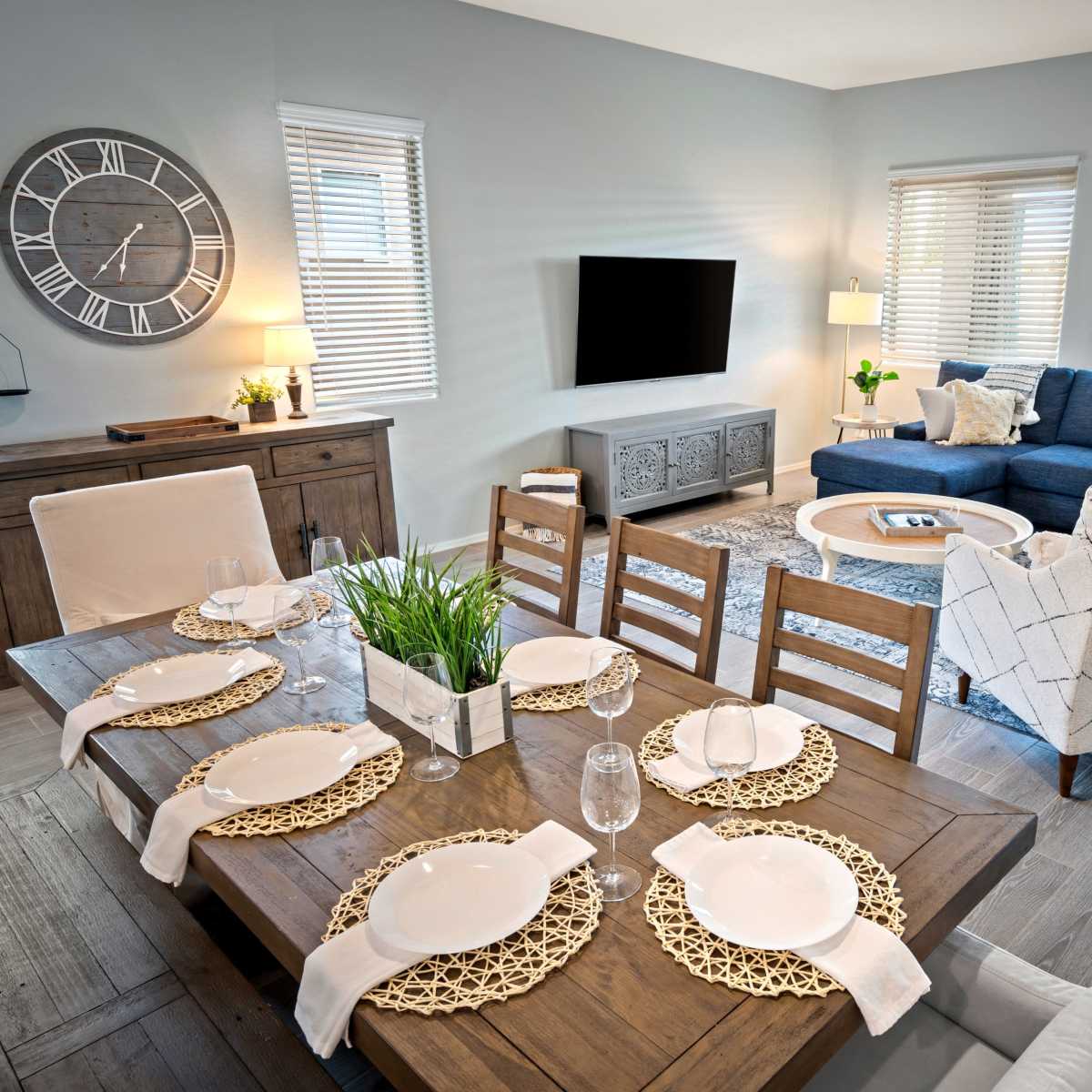 A modern living and dining area featuring a wooden dining table set for six, a dark blue sectional couch, and a wall-mounted TV in a light-colored room with natural light from several windows.