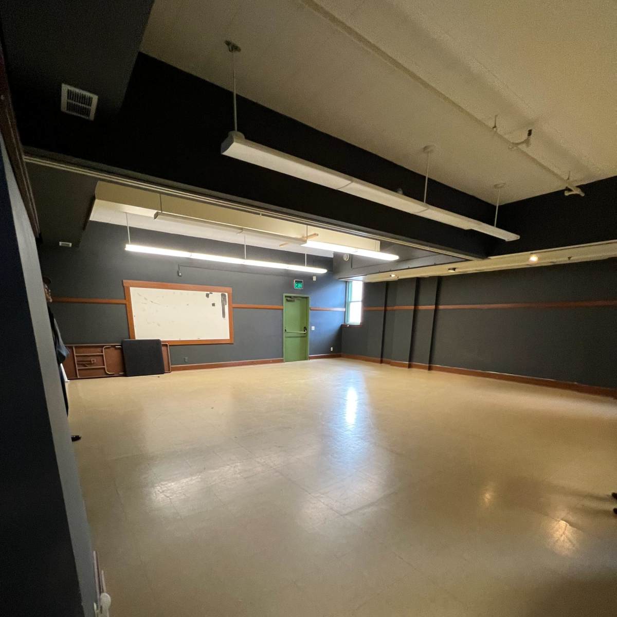 An empty room with light-colored flooring, dark walls, and a green door, featuring overhead lighting and a whiteboard on one wall.