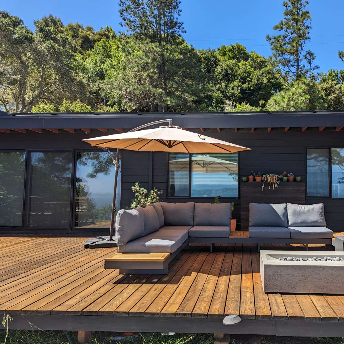 A wooden deck features a sectional sofa, a large umbrella, and a fire pit in front of a house surrounded by trees and a view of the water.