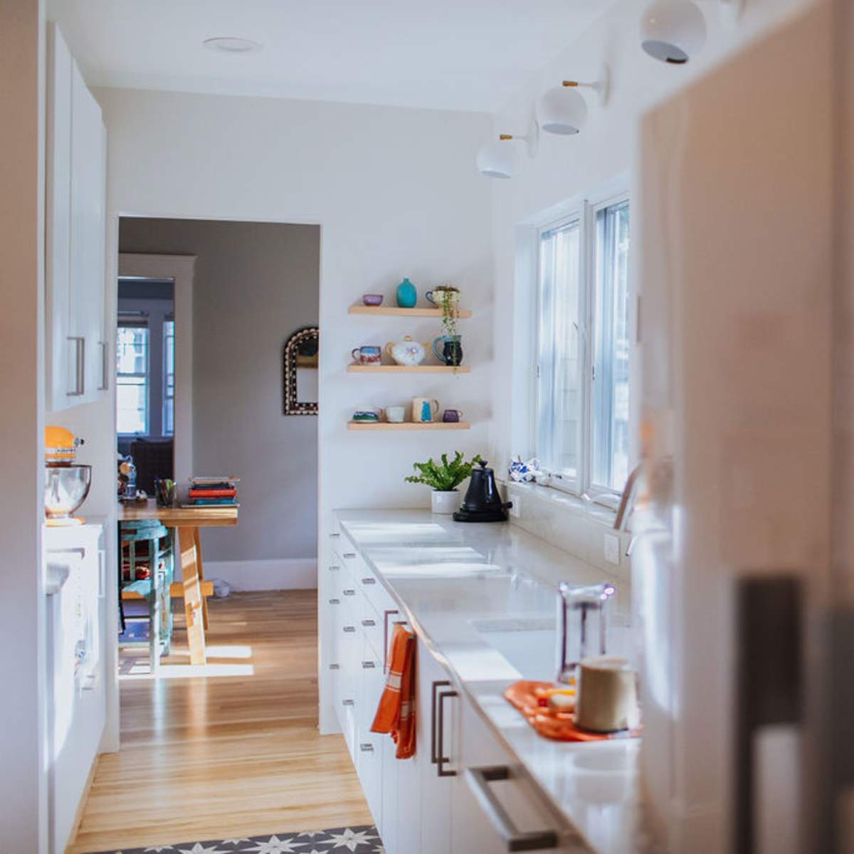 A modern kitchen with white cabinetry, a view into a dining area with colorful tableware on shelves, and natural light coming through large windows.