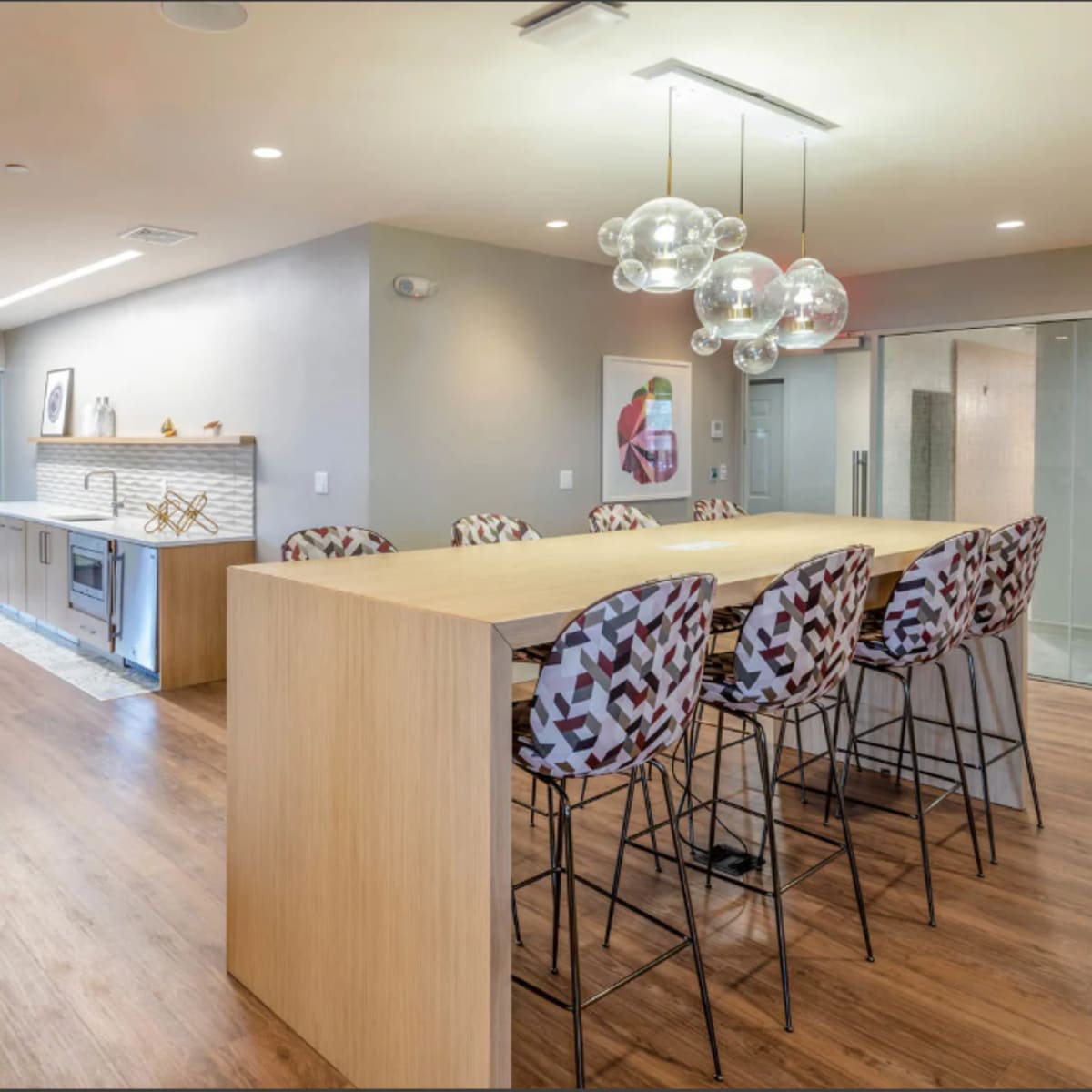 A modern kitchen and dining area with a long wooden bar surrounded by stylish high chairs, and a living area featuring a cozy chair.
