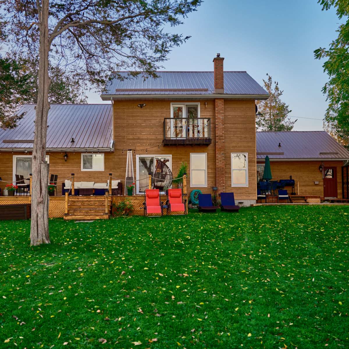 A two-story wooden house with a metal roof is situated in a grassy area surrounded by trees.