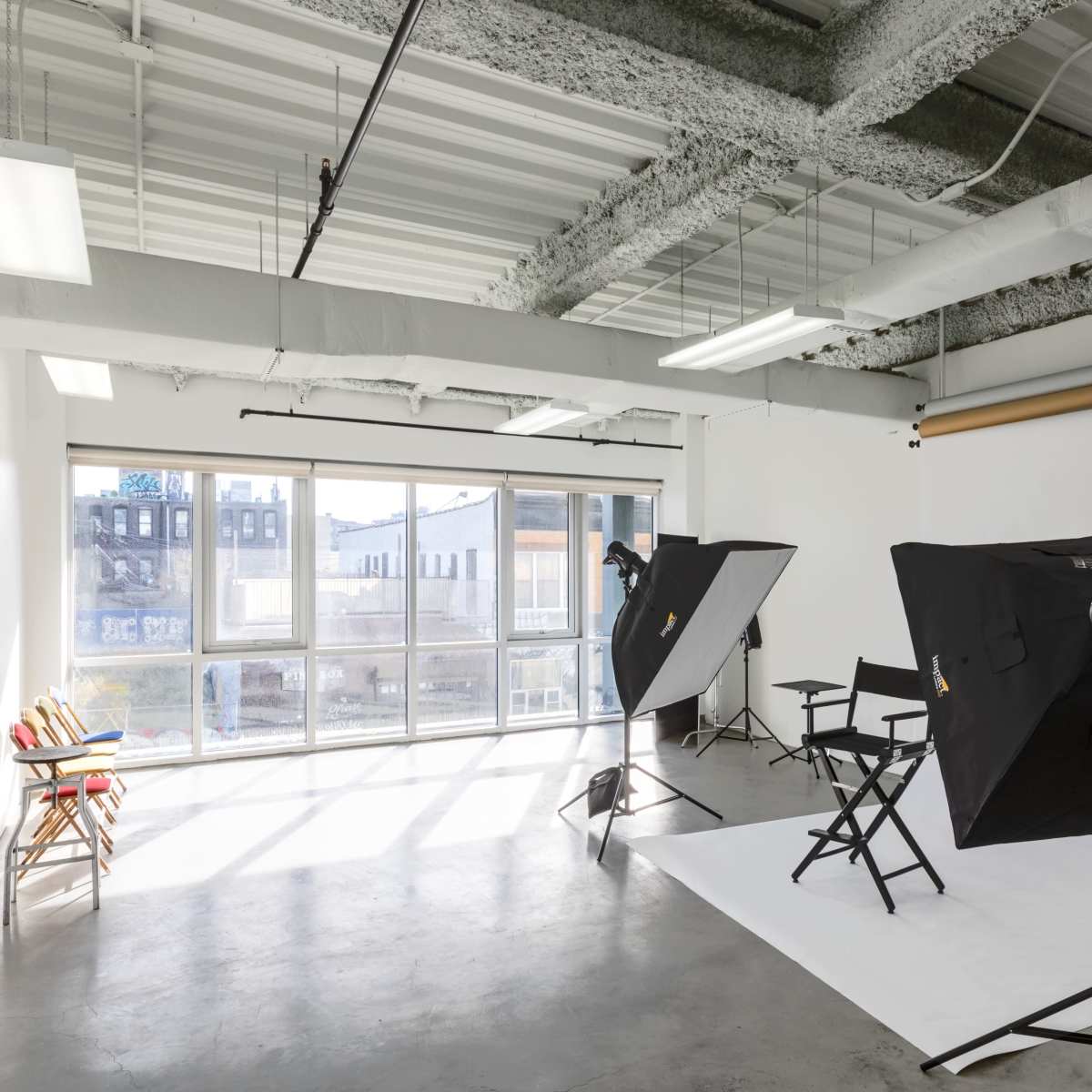 A photography studio equipped with two lighting setups, a backdrop, and a director's chair near large windows that allow natural light.