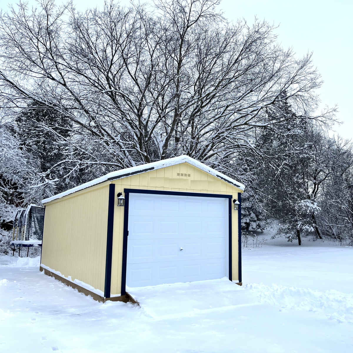 A yellow shed with a white garage door is surrounded by a snowy landscape and bare trees.
