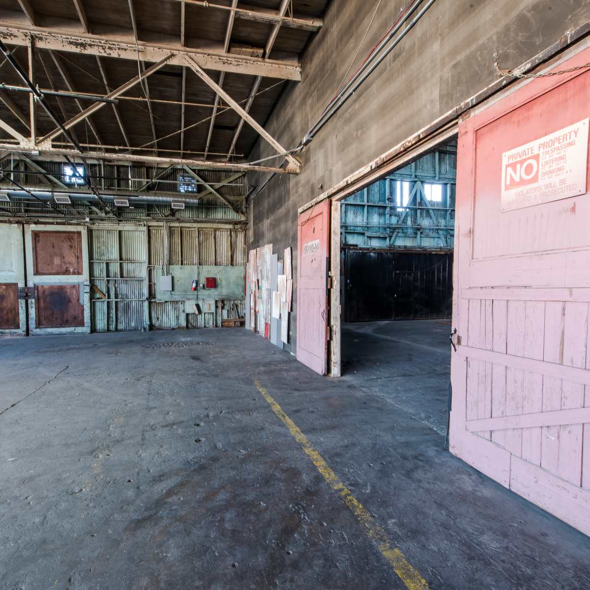The interior of a large, empty warehouse featuring open doors, exposed beams, and a concrete floor.