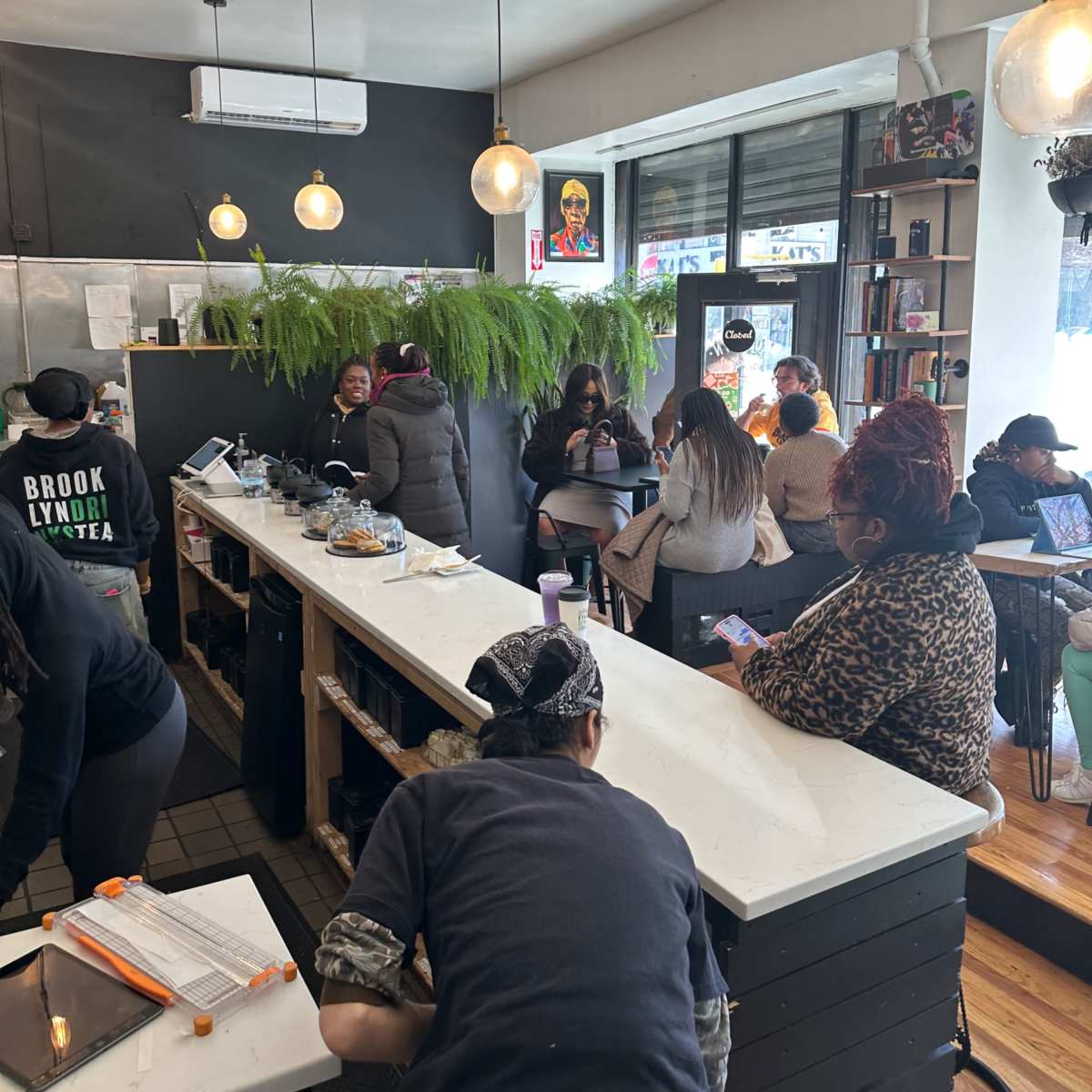A busy cafe with customers seated, employees preparing food, and a large plant decoration along the counter.