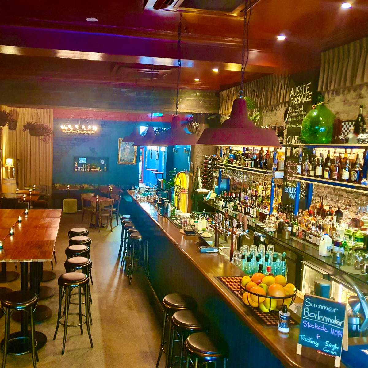 A dimly lit bar with a long counter, high stools, a wooden communal table, and various bottles arranged on shelves behind the bar.