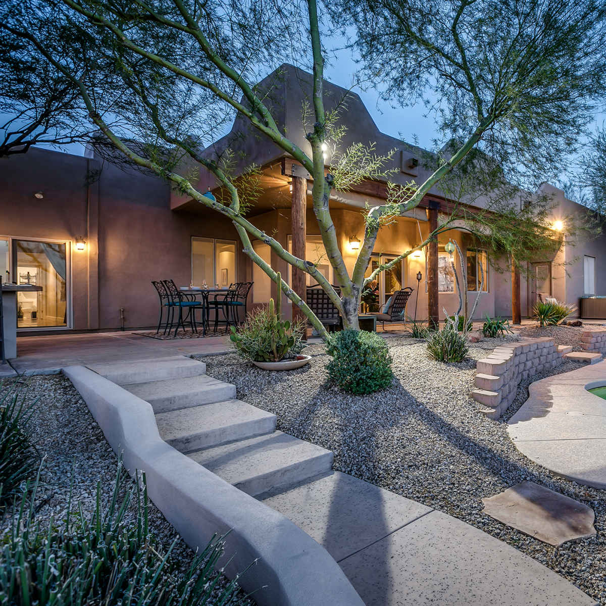 A home with a backyard featuring a swimming pool, landscaped gardens, and patio seating under tree branches at dusk.