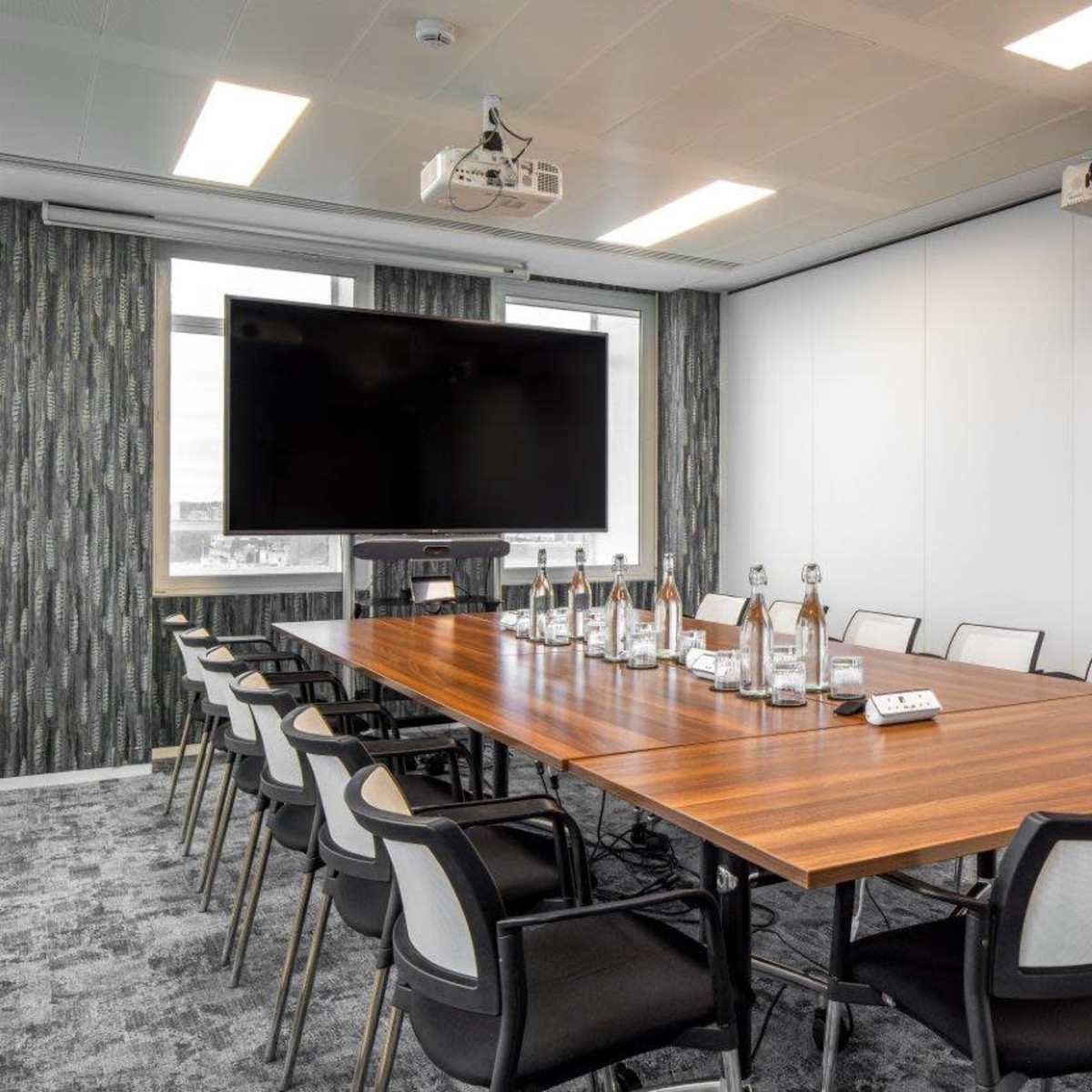A modern conference room features a long wooden table surrounded by chairs, with a large screen and projector mounted on the wall.