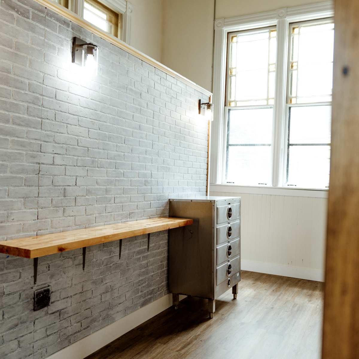A room with a wooden counter along a gray brick wall, alongside a metal drawer unit, illuminated by wall sconces and natural light from large windows.