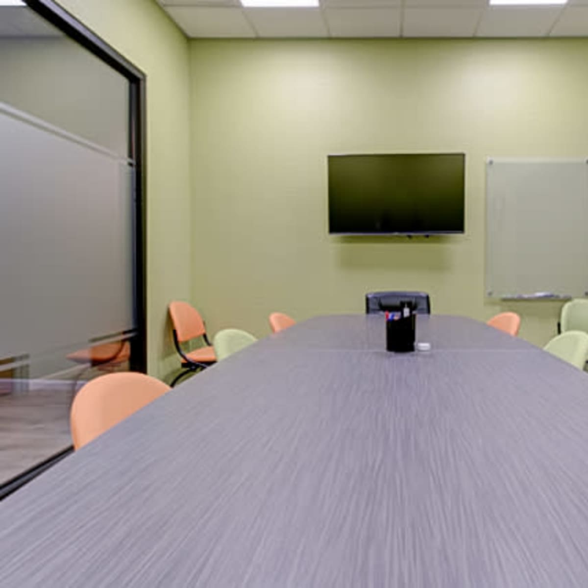 A conference room features a long gray table with orange chairs, a television mounted on the wall, and large glass panels.