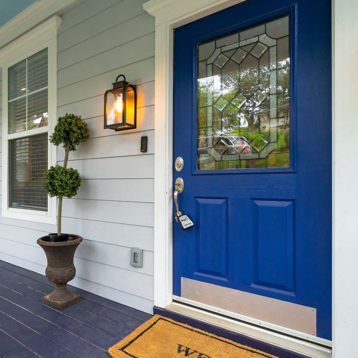 A front porch with a blue door, a potted plant, a chair, and a welcome mat on a patterned rug.