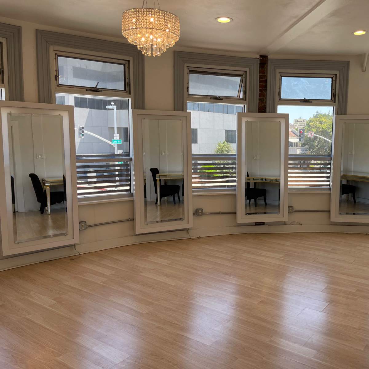 A sunlit room with wooden flooring features five tall mirrors along the curved wall and a table with a chair in the corner.