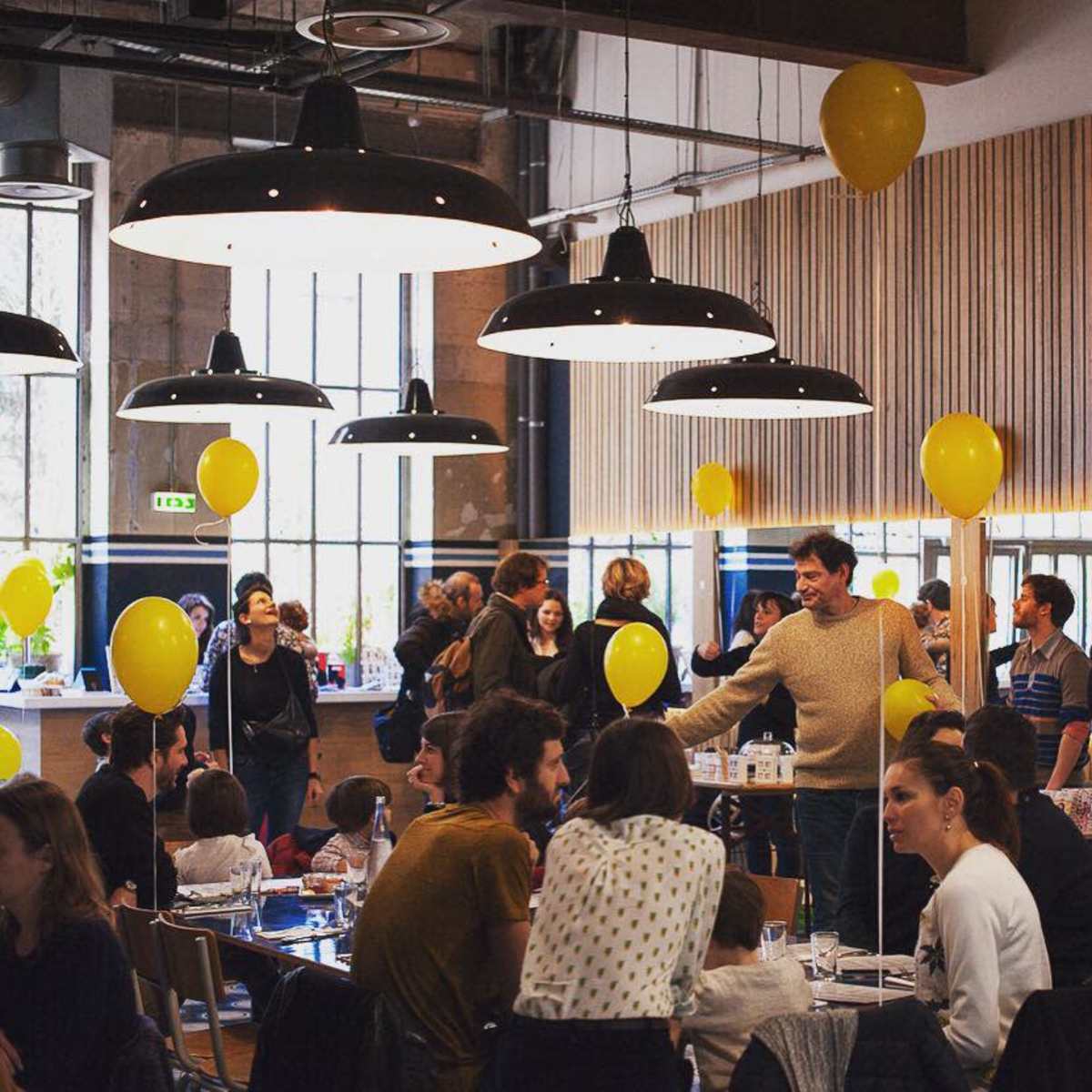 A lively gathering takes place in a spacious venue filled with people, yellow balloons, and modern lighting fixtures.