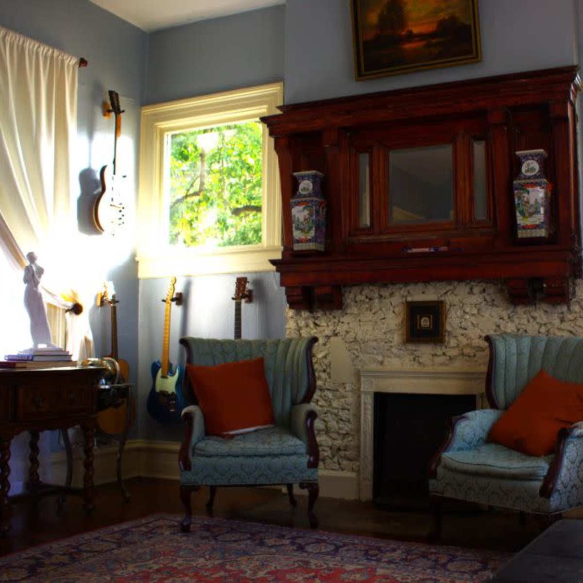 A cozy living room with two patterned armchairs, a wooden fireplace surrounded by stone, a small table, and various guitars displayed on the walls.