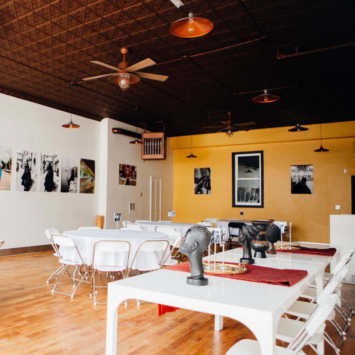 A spacious, well-lit restaurant interior with several tables set up, wall art featuring black and white photographs, and ceiling fans.