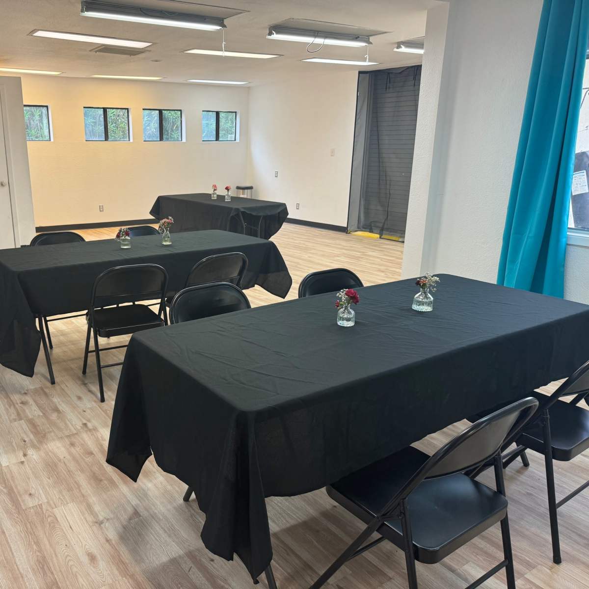 A room set up with several black tables, each adorned with small flower vases, and a few windows allowing light to enter.