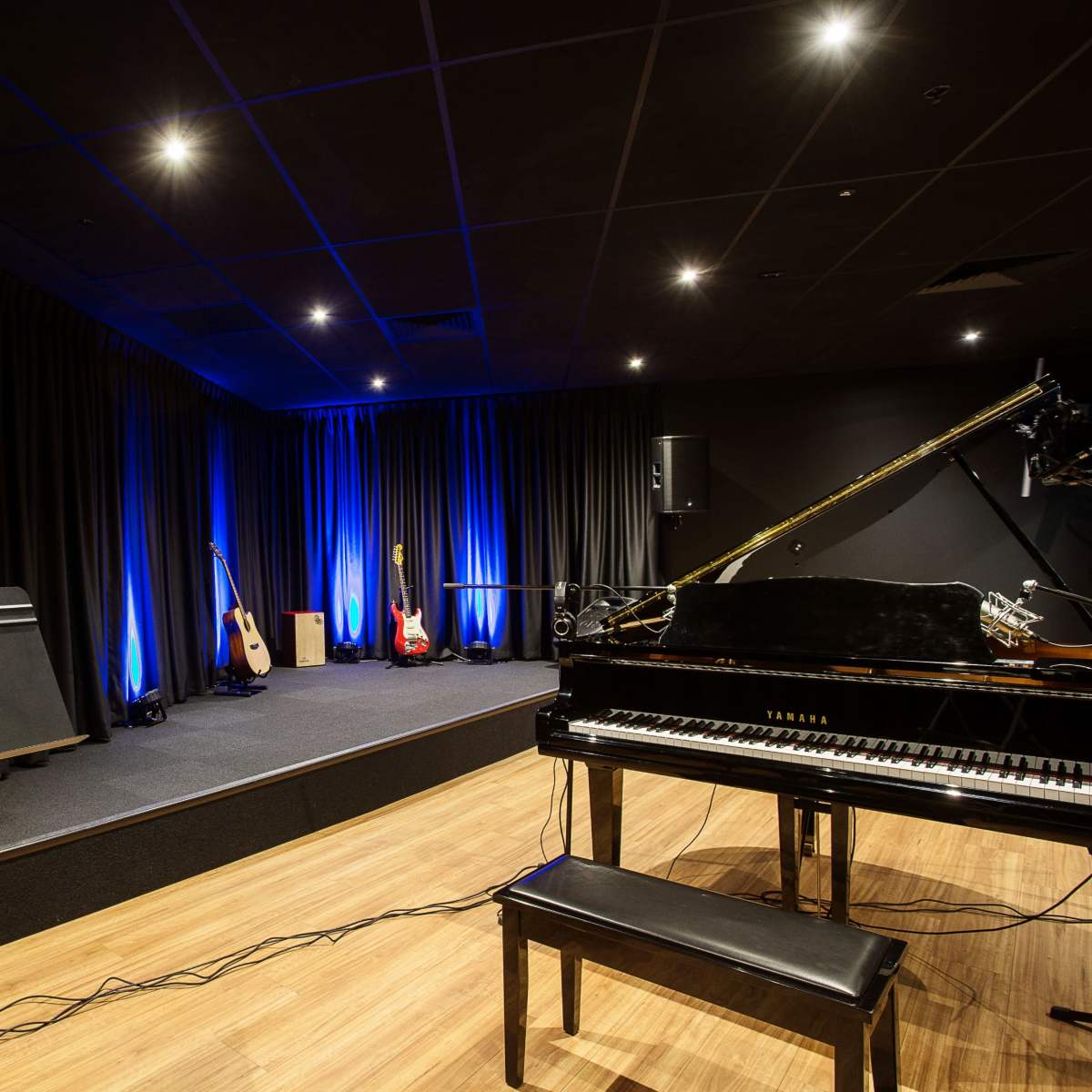 A music studio featuring a grand piano, microphones, audio equipment, and illuminated blue lights against a black curtain backdrop.