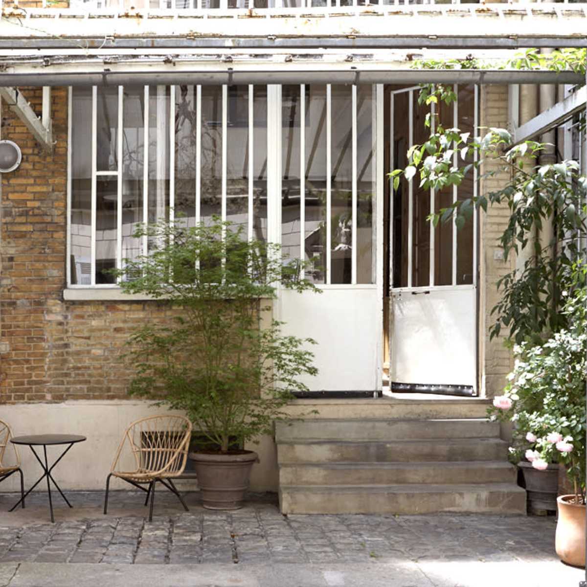 A small outdoor seating area features two chairs and a table in front of a brick wall with large windows and greenery.