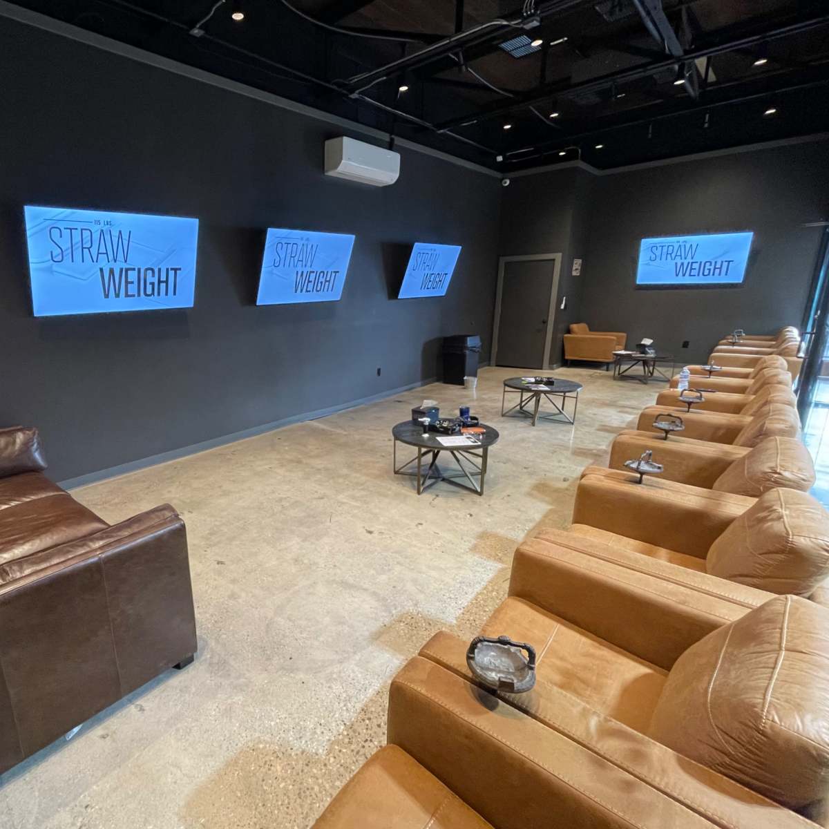 A modern waiting area features brown leather seating, large screens displaying "STRAW WEIGHT," and minimalist decor.