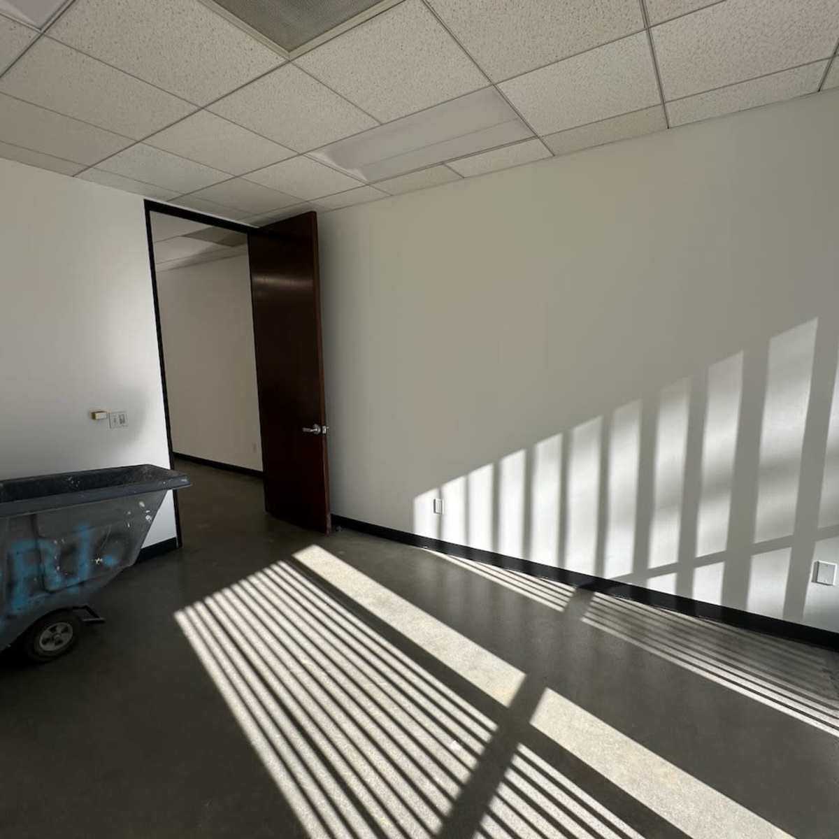 A dimly lit room features a doorway, a wheelbarrow in the corner, and shadows cast by vertical blinds on the concrete floor.