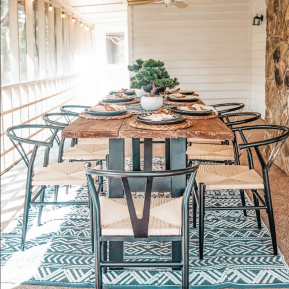 A long dining table is set with plates and decorative items, surrounded by chairs, in a sunlit porch with wooden walls and a stone accent.