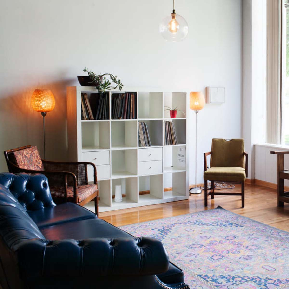 A cozy living room with a blue leather sofa, a wooden bench, a bookshelf filled with records, and large windows allowing natural light to enter.
