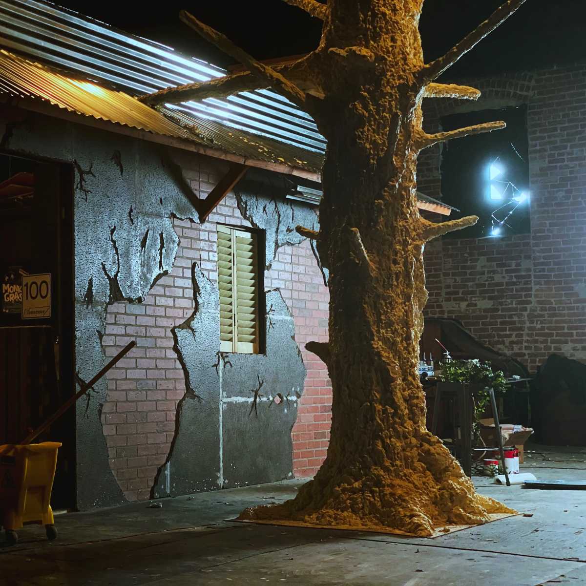 A dimly lit outdoor scene featuring a textured wall with cracks and a large, rough-textured tree structure next to a yellow mop bucket.