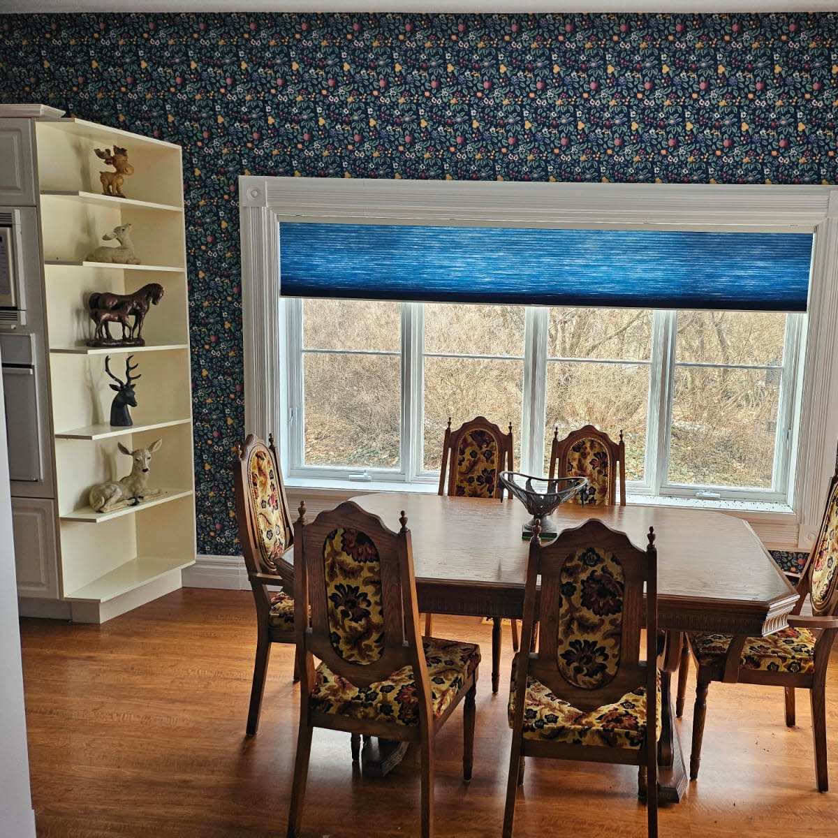 A dining room features a wooden table surrounded by ornate chairs, with large windows offering a view of a landscape beyond a floral wallpapered wall.