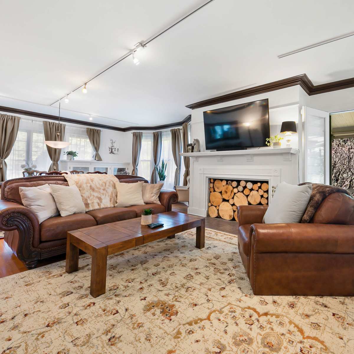 The living room features two leather sofas, a wooden coffee table, and a fireplace with stacked firewood, leading to a sunlit porch with large doors.