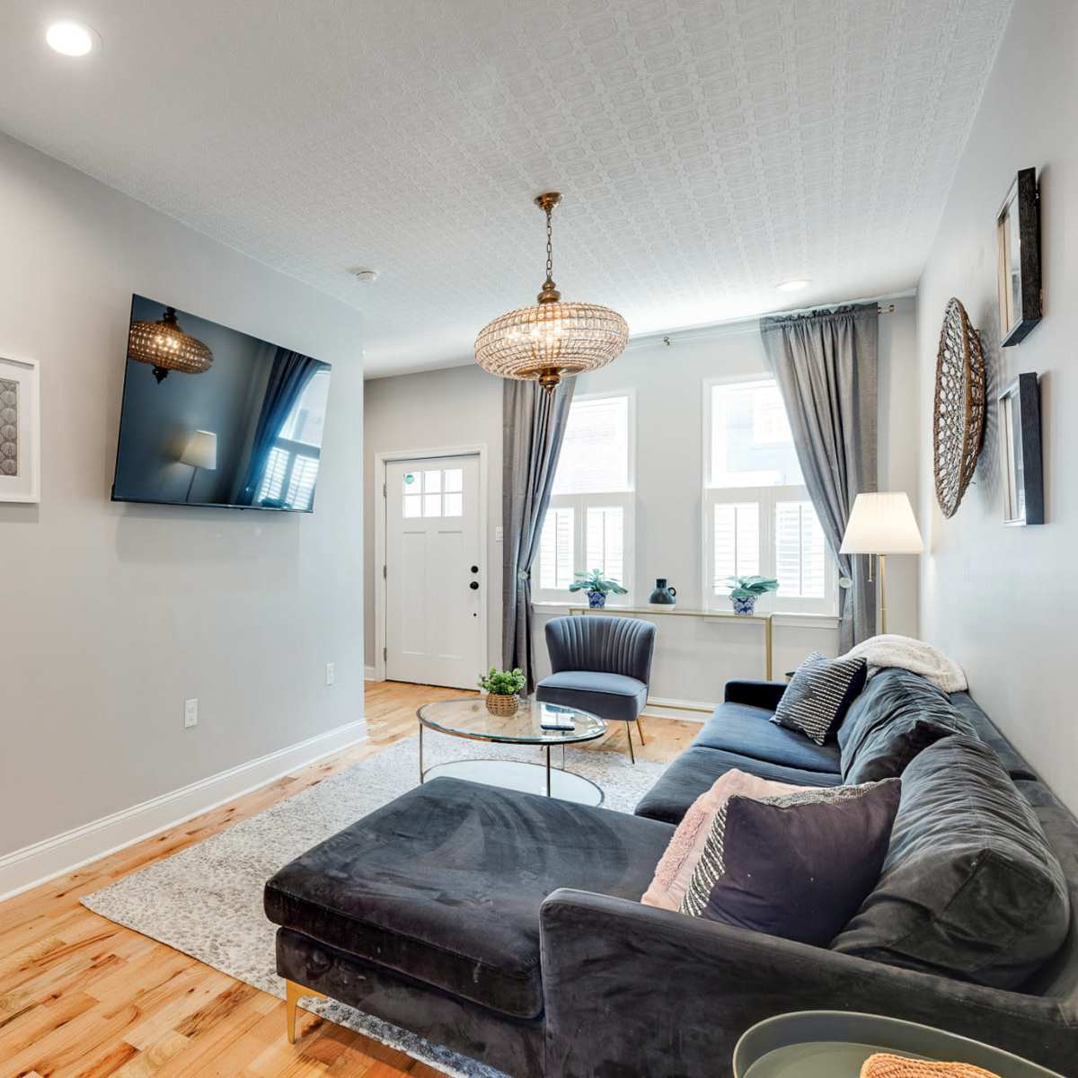 The living room features a dark sofa, a flat-screen TV mounted on the wall, and large windows with light drapes.